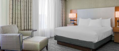 Habitación de hotel con una cama grande, una silla, un reposapiés, una mesita y una lámpara.