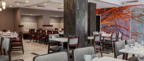 Un restaurante moderno con techos altos, iluminación empotrada y un amplio mural abstracto. Las mesas están dispuestas con manteles blancos y cubiertos.