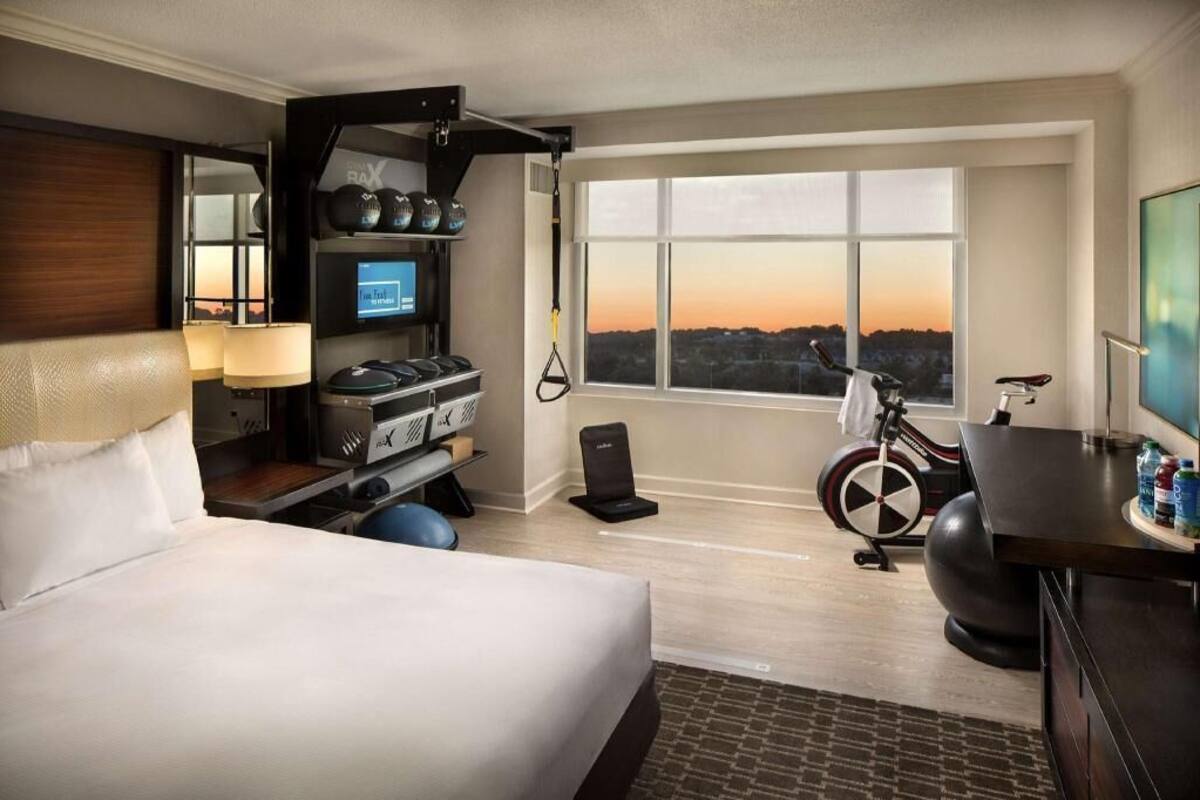 A hotel room with a bed, exercise equipment, a TV, and a large window with a sunset view.