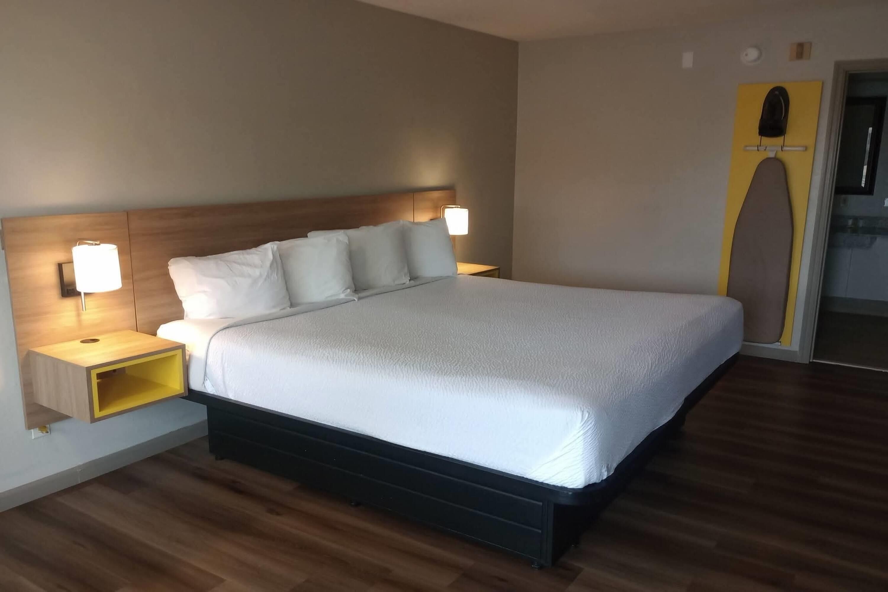 A modern hotel room with a large bed, bedside tables with lamps, a wooden headboard, and a built-in ironing board.