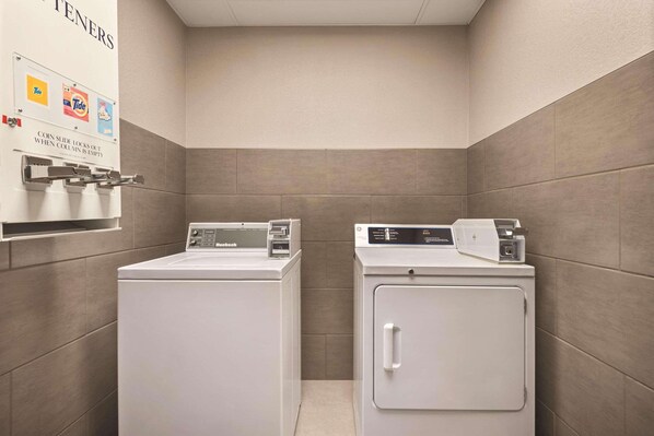 A laundry room with two front-loading washers and dryers, a coin-operated dryer, and a wall-mounted sign with detergent brands.