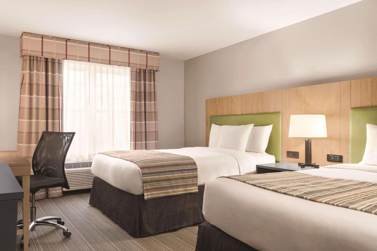 A hotel room with two beds, a desk, a chair, and a lamp.