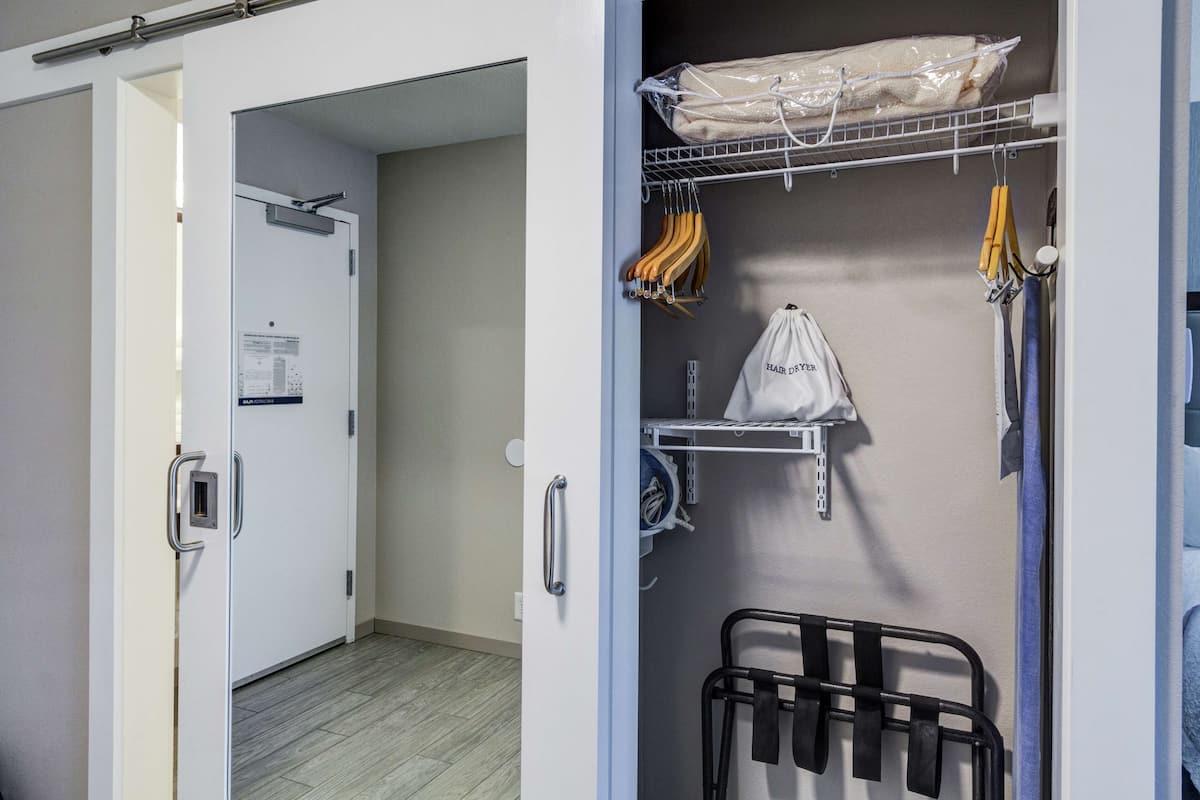 Une chambre d’hôtel dotée d’un dressing où sont rangés des vêtements sur des cintres, une étagère et une chaise noire.