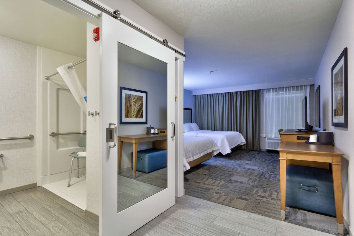 Une chambre d’hôtel comprenant un lit, un bureau, une chaise, une petite table, un miroir et un espace douche.