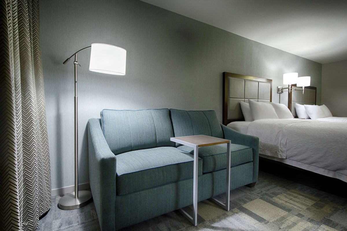 Une chambre d’hôtel avec un canapé vert, une petite table, un lit avec une literie blanche et une lampe sur pied.