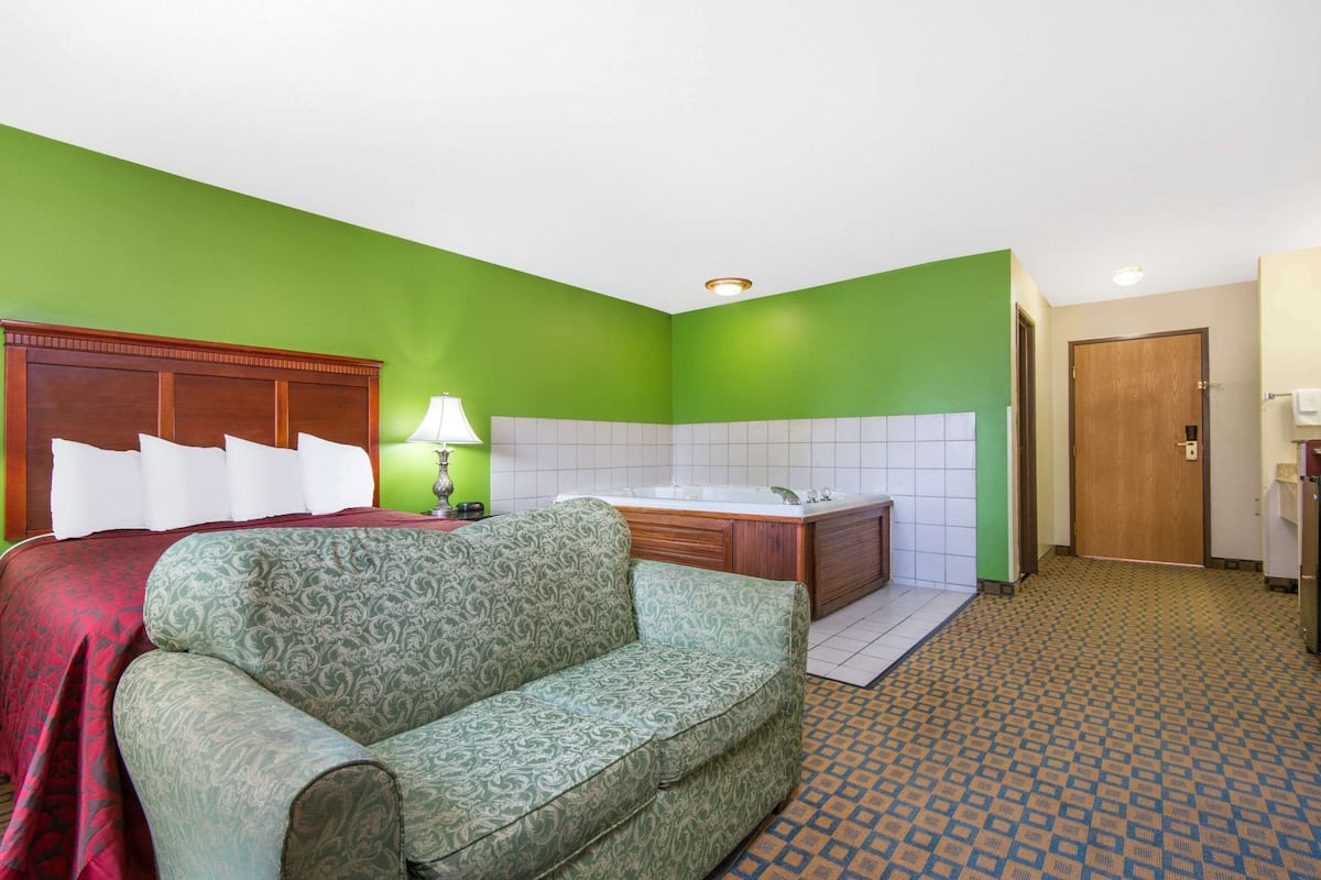 A hotel room with a green wall, a large bed with white pillows, a green patterned sofa, a bathroom with a bathtub, and a patterned floor.