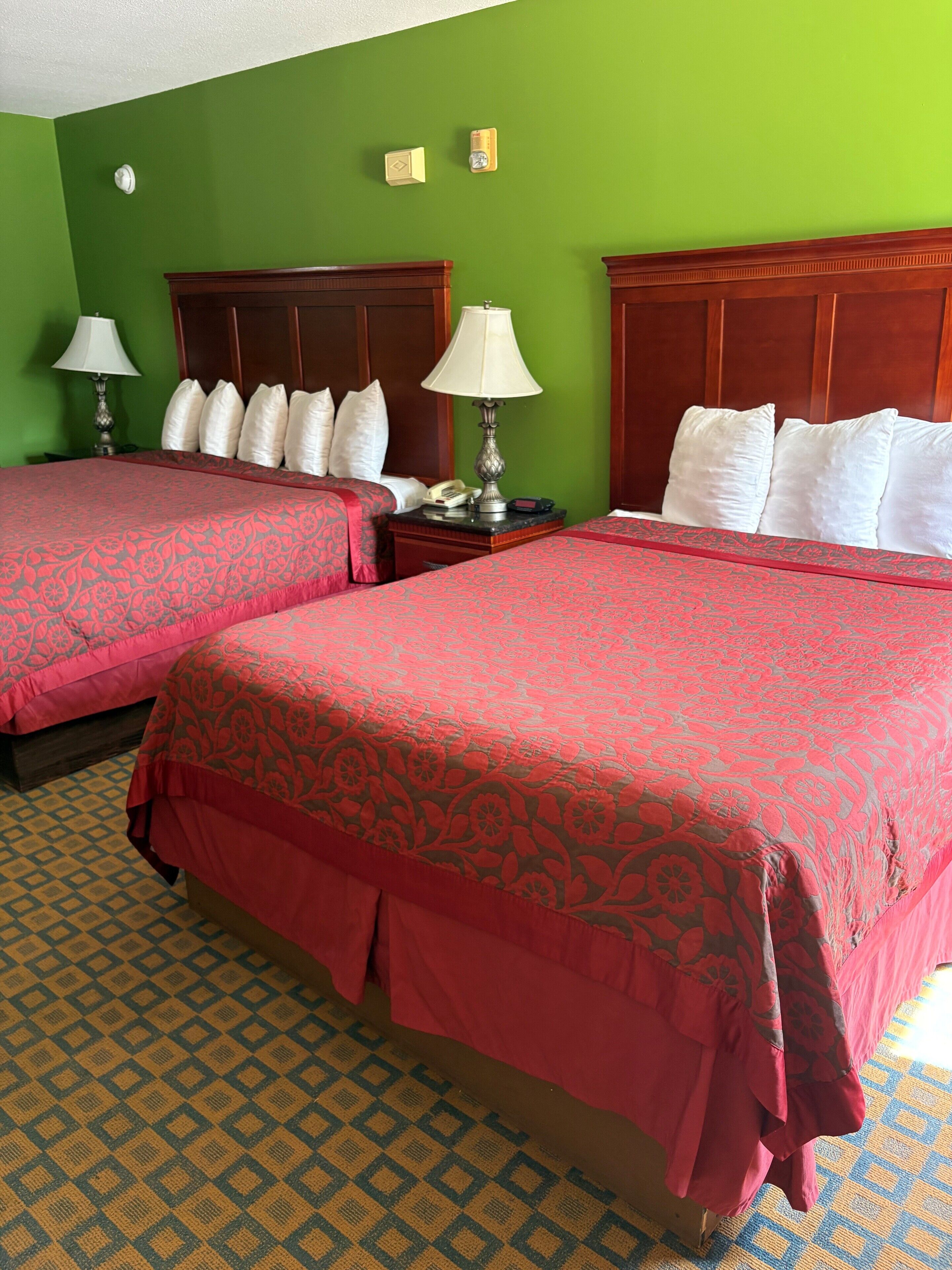 Habitación de hotel con dos camas, paredes verdes, colchas rojas estampadas, cabeceros de madera, lámparas de noche y piso con diseño.