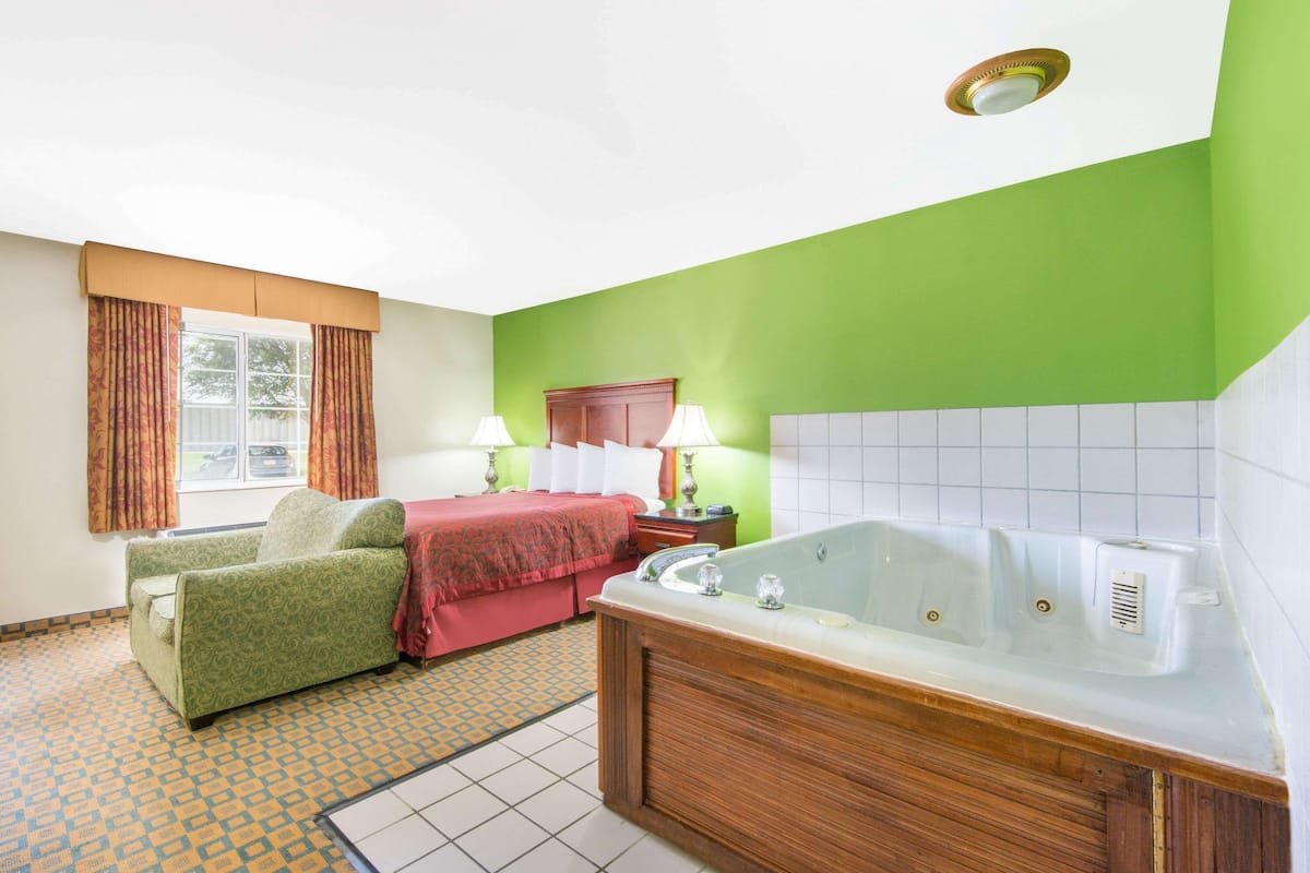 A hotel room with a green wall, a large bathtub, a sofa, a bed with a red blanket, and a window with curtains.
