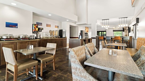 A modern hotel dining area with high tables and comfortable seating, a kitchen area with a coffee machine, and a large window providing a view of the outdoors. - Best Western Paducah Inn (Paducah)