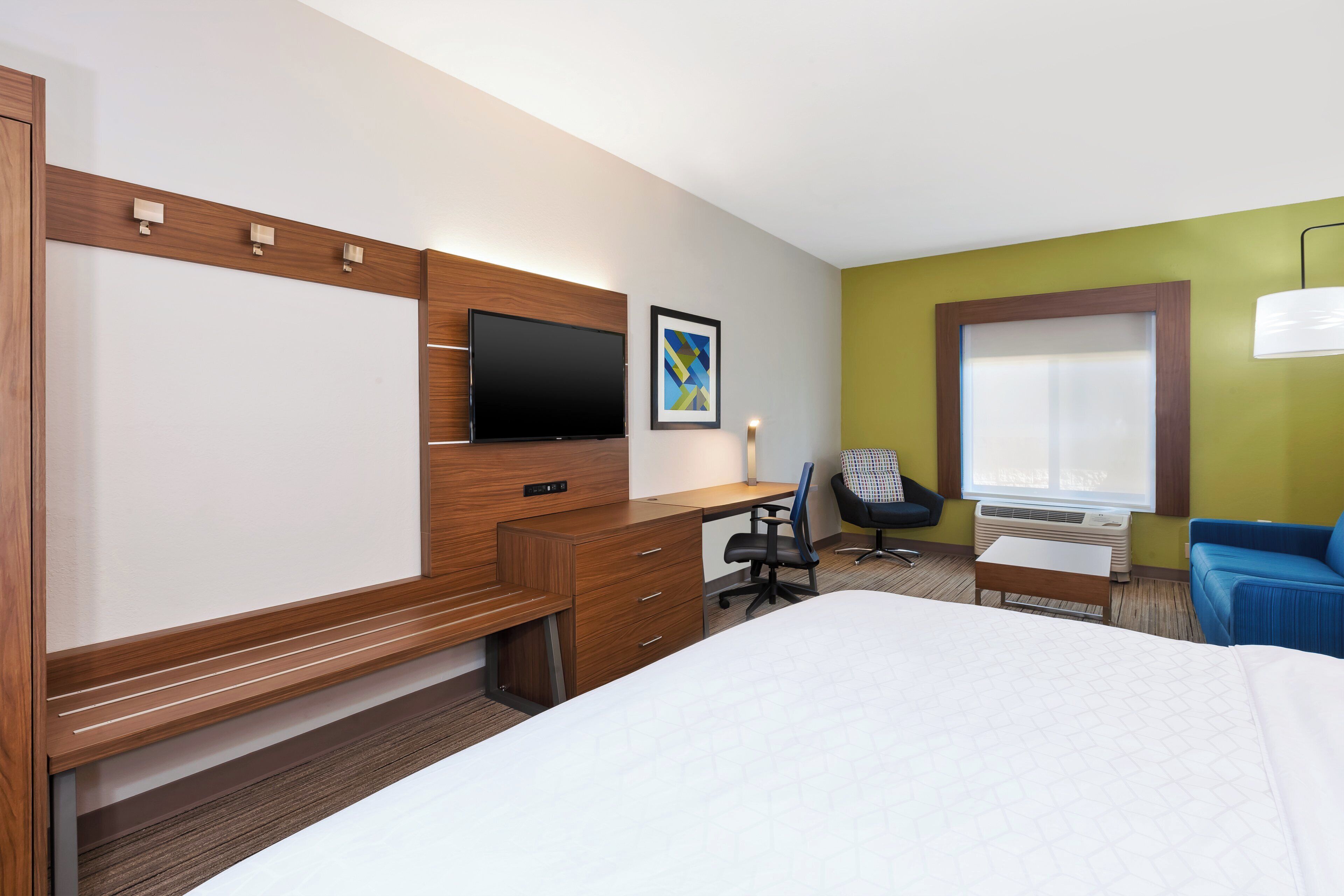 A hotel room with a bed, desk, chair, TV, and air conditioning unit.