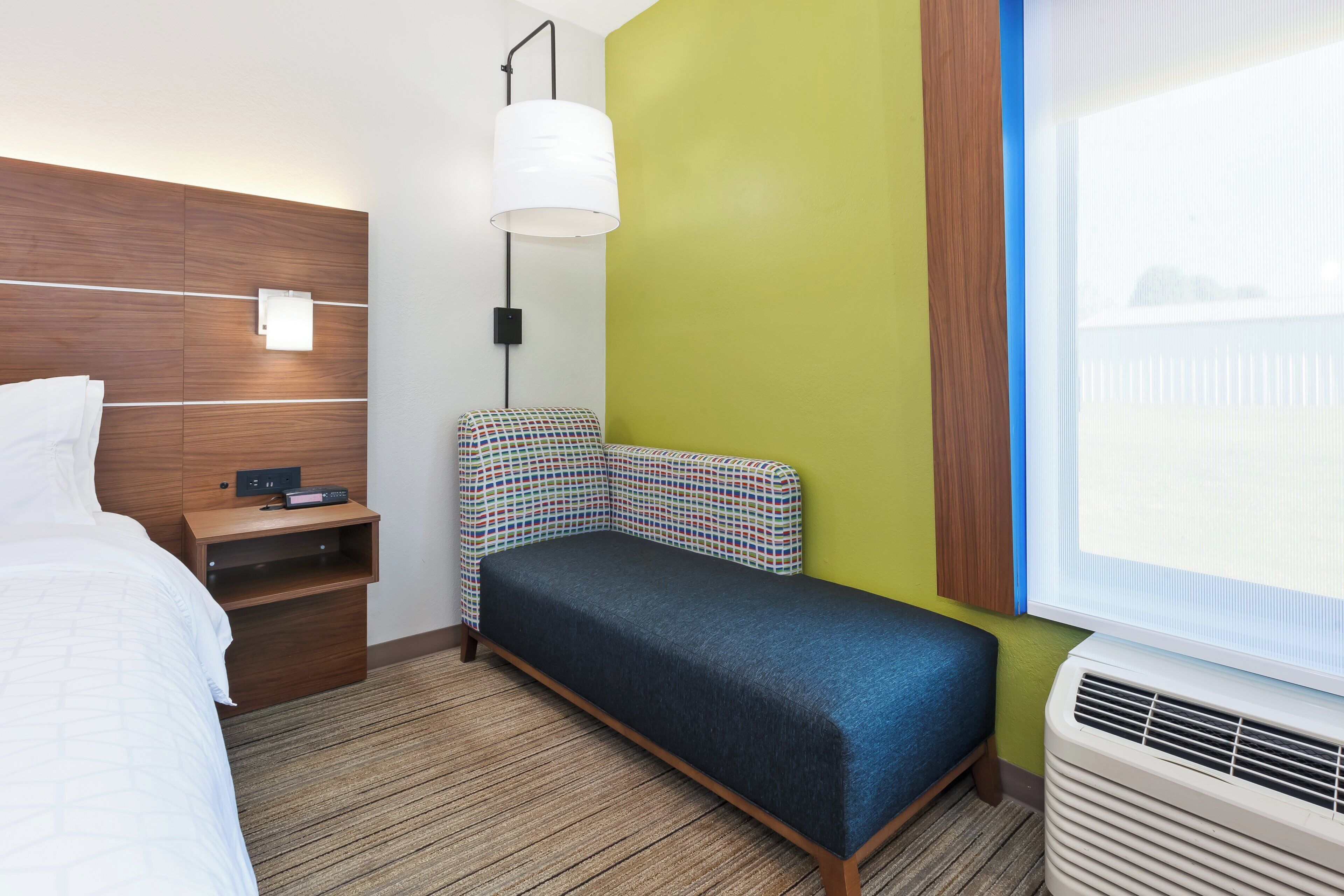 Habitación de hotel con cama, mesita de noche con radio, lámpara de pared y ventana con persianas.