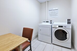 A guest laundry area with a washing machine, dryer, and a table with a chair. A sign above the machines states 'Guest Laundry' with usage instructions. - Holiday Inn Express & Suites Three Rivers by IHG (Three Rivers)