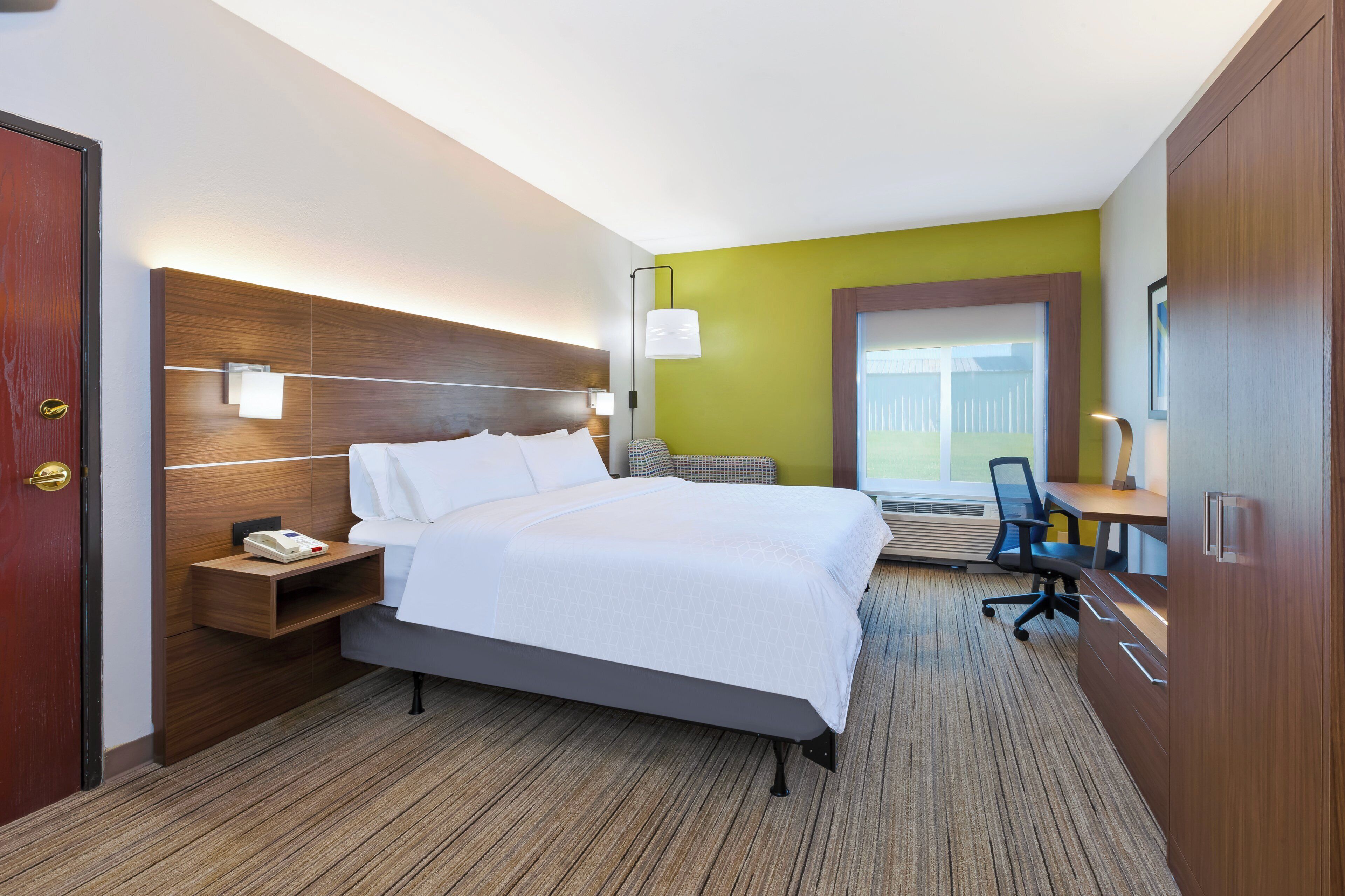 A hotel room with a bed, bedside table, lamp, desk, chair, and a view of the outside.