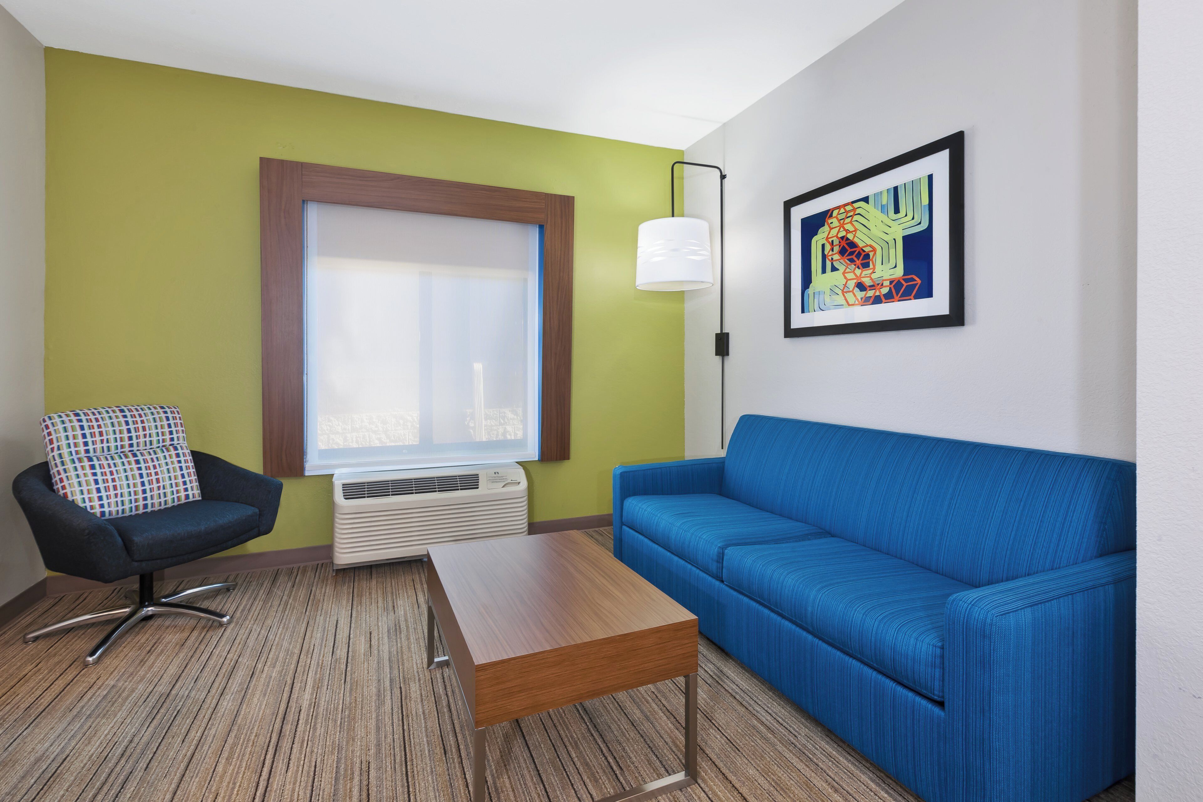 A room with a blue sofa, a black chair with a colorful cushion, a wooden coffee table, a window with blinds, a wall-mounted lamp, and a framed abstract painting.