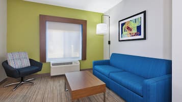 A room with a blue sofa, a black chair with a colorful cushion, a wooden coffee table, a window with blinds, a wall-mounted lamp, and a framed abstract painting.
