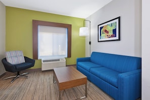 Quarto com sofá azul, cadeira preta com almofada colorida, mesa de centro de madeira, janela com persianas, luminária fixada na parede e quadro abstrato emoldurado.
