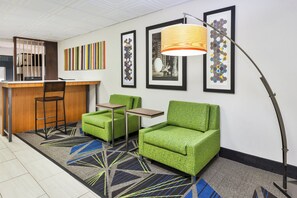 A hotel lobby with a reception desk, two green armchairs, a small table, a floor lamp, and abstract art on the walls.