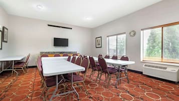 Un salón de conferencias con mesas y sillas, un televisor de pantalla plana, cuadros enmarcados y ventanas amplias con persianas.