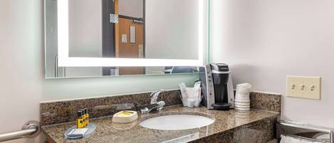 Baño de hotel con lavamanos de encimera de granito, un espejo grande, cafetera y un perchero para toallas.