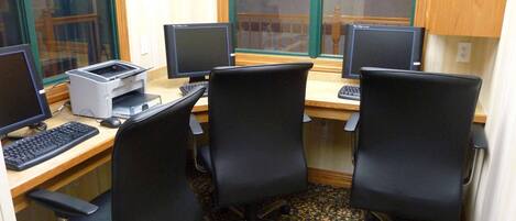 Um laboratório de informática com três mesas, cada uma com monitor, teclado e impressora. Cadeiras dispostas ao redor das mesas.