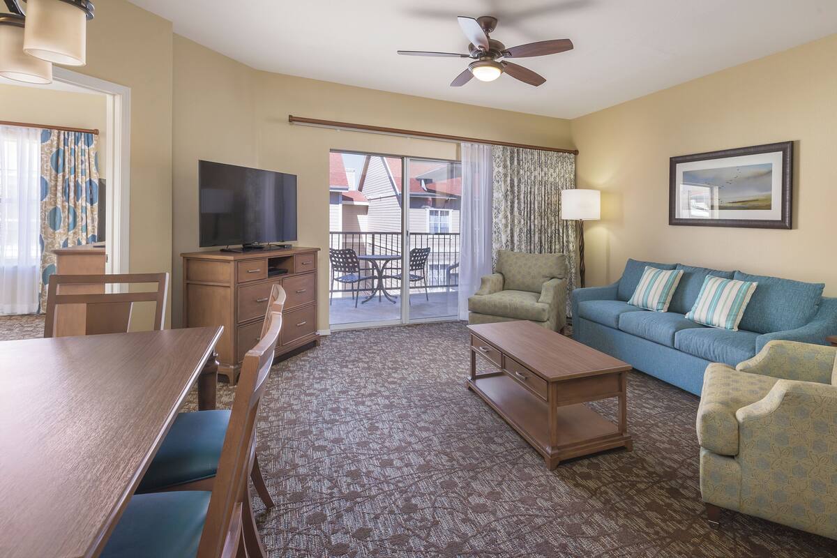 Habitación de hotel con televisor, sofá, sillón, mesa de comedor y balcón con mobiliario exterior.