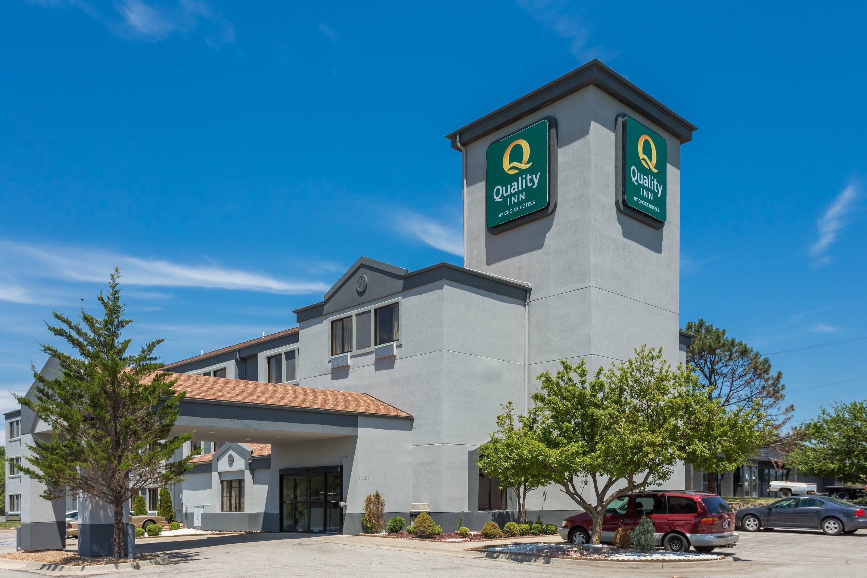 Hotel Quality Inn com uma torre imponente exibindo o logotipo da Quality Inn, fachada de edifício moderna e estacionamento com vários carros.
