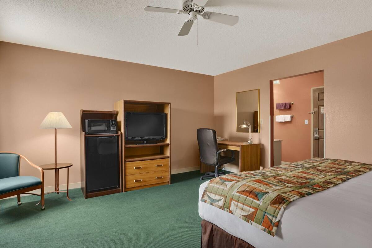 A hotel room with a bed, a desk with a chair, a television, a microwave, a refrigerator, and a ceiling fan.