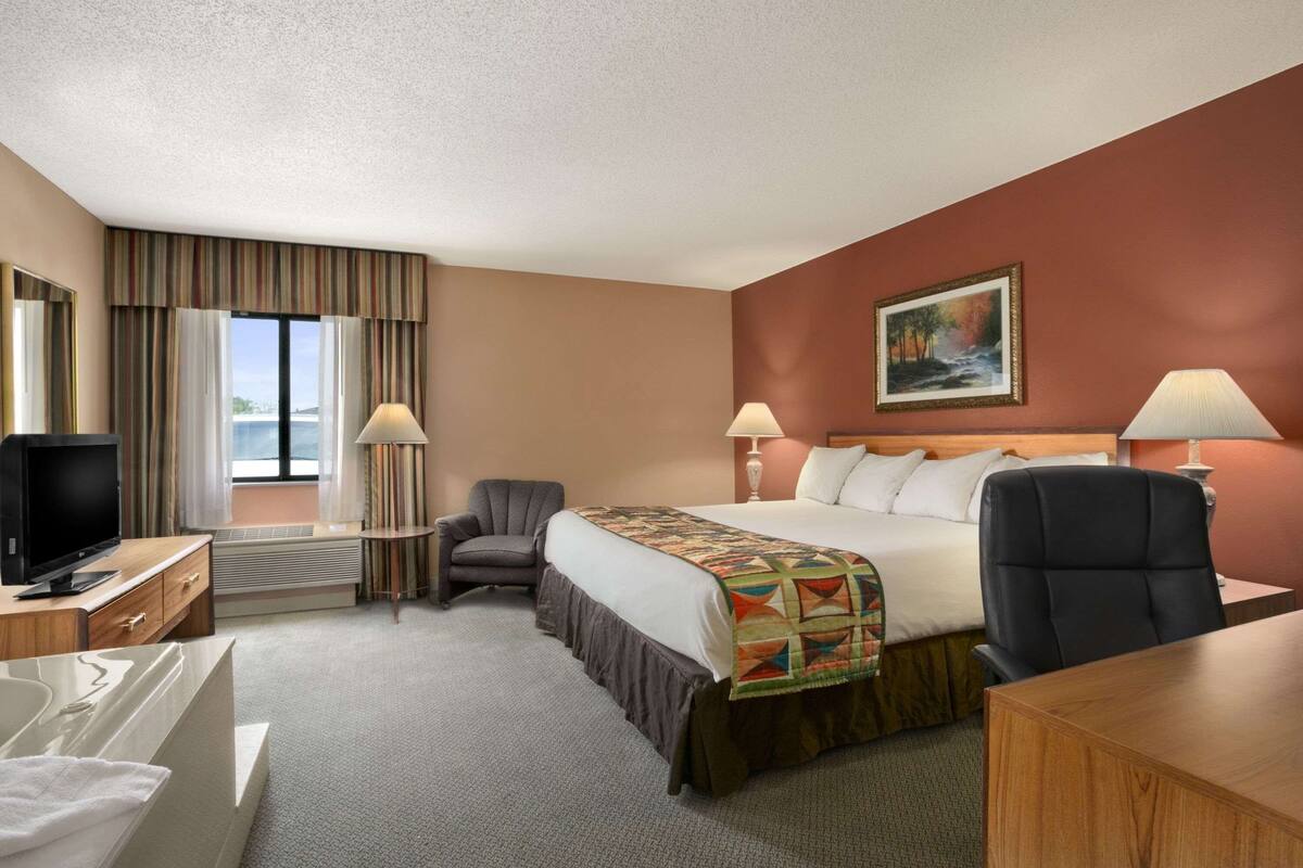 A hotel room with a bed, two chairs, a desk, a TV, and a painting on the wall.