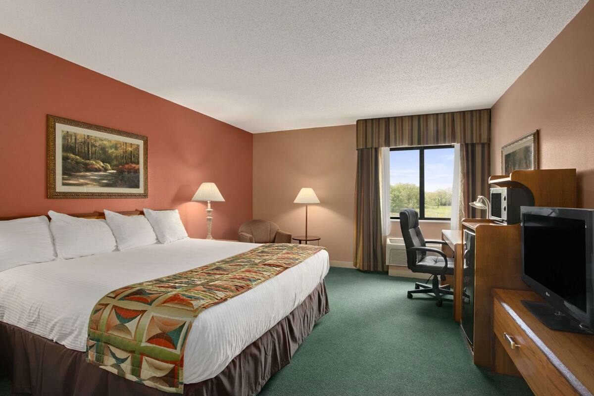 A hotel room with a large bed, a desk with a television, a chair, and a window with a view of trees.