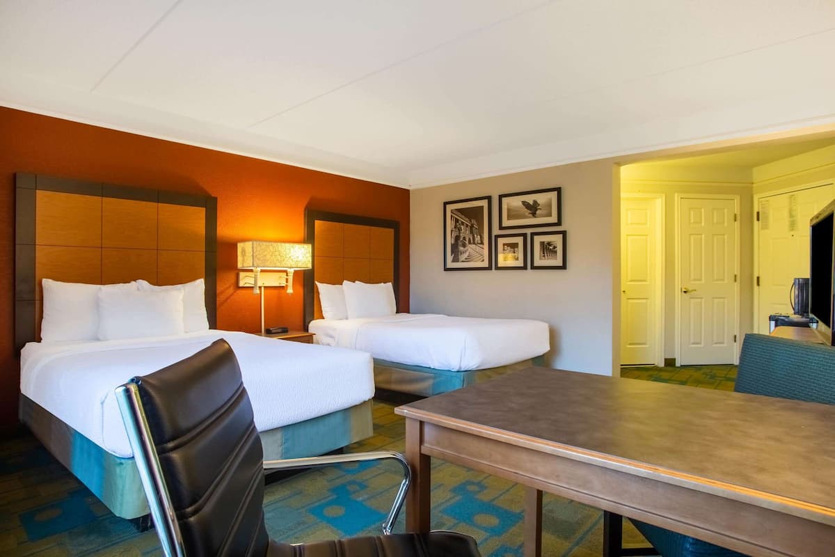 A hotel room with two beds, a desk, and a chair. There are framed pictures on the wall and a door leading to another room.