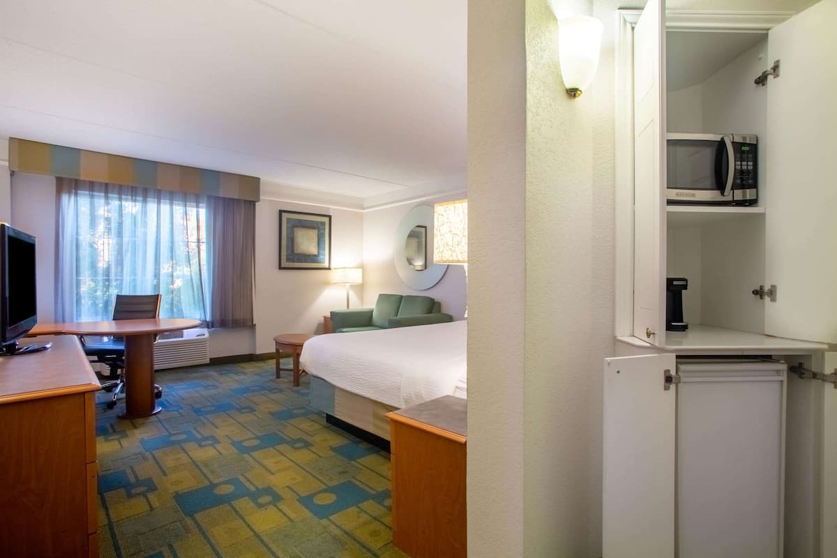 A hotel room with a bed, a desk with a computer, a chair, a lamp, a microwave, and a small refrigerator.