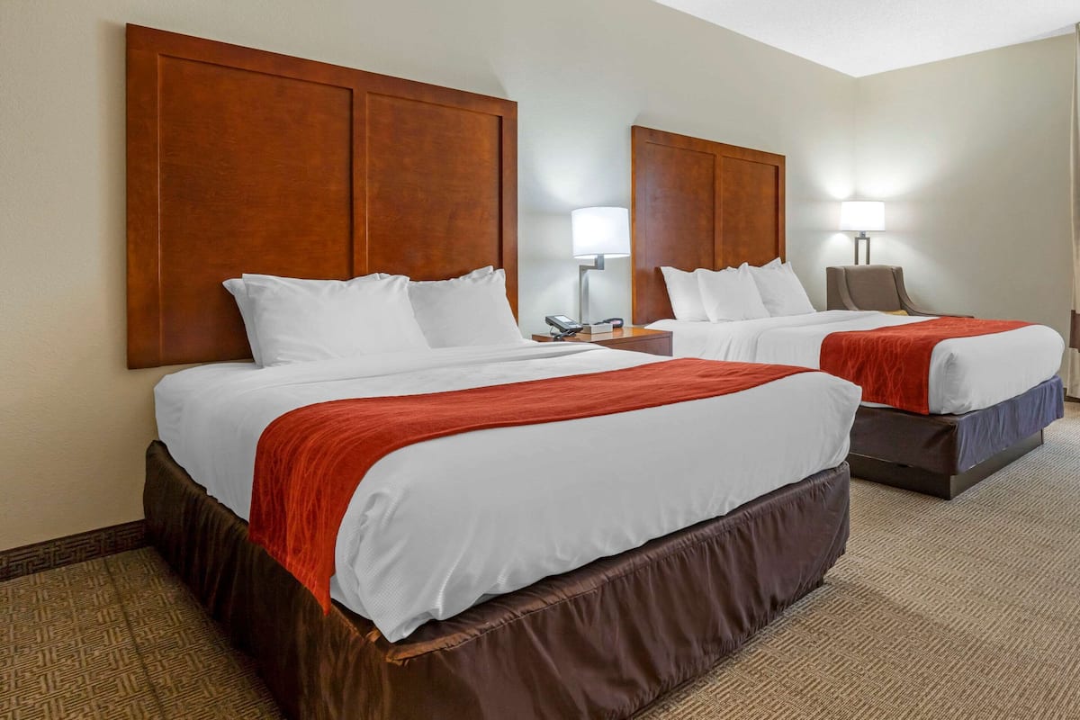 A hotel room with two beds, each with a wooden headboard, white bedding, and a red throw blanket. There are bedside tables with lamps and a small table between the beds.