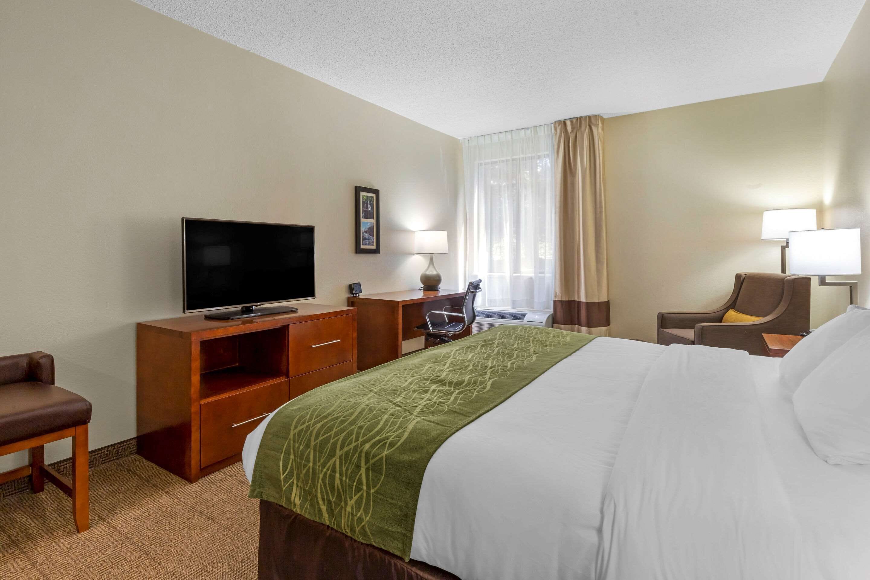 A hotel room with a large bed, a television on a wooden dresser, a desk with a chair, and a window with curtains.