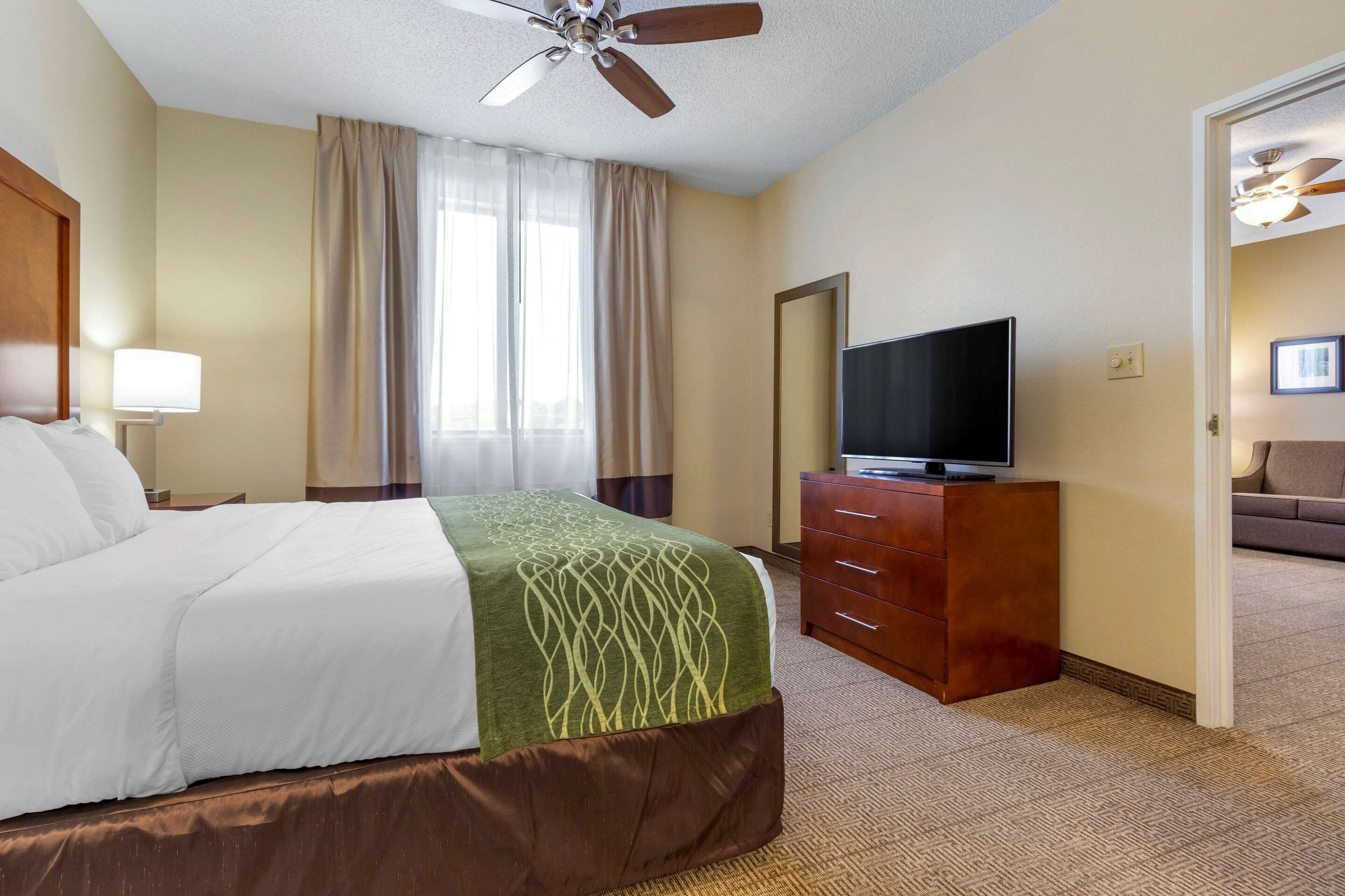 A hotel room with a bed, a television on a wooden dresser, a ceiling fan, and a view of a hallway with a couch.