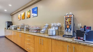 Zona de desayuno autoservicio con máquina de café, dispensadores de alimentos y televisor montado en la pared.