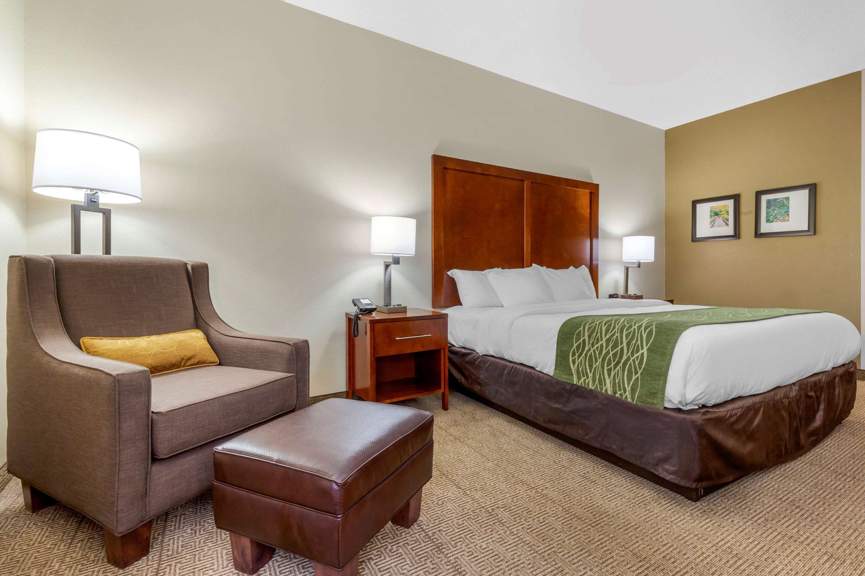 Una habitación de hotel con una cama, un sillón, un reposapiés y dos mesitas de noche con lámparas.