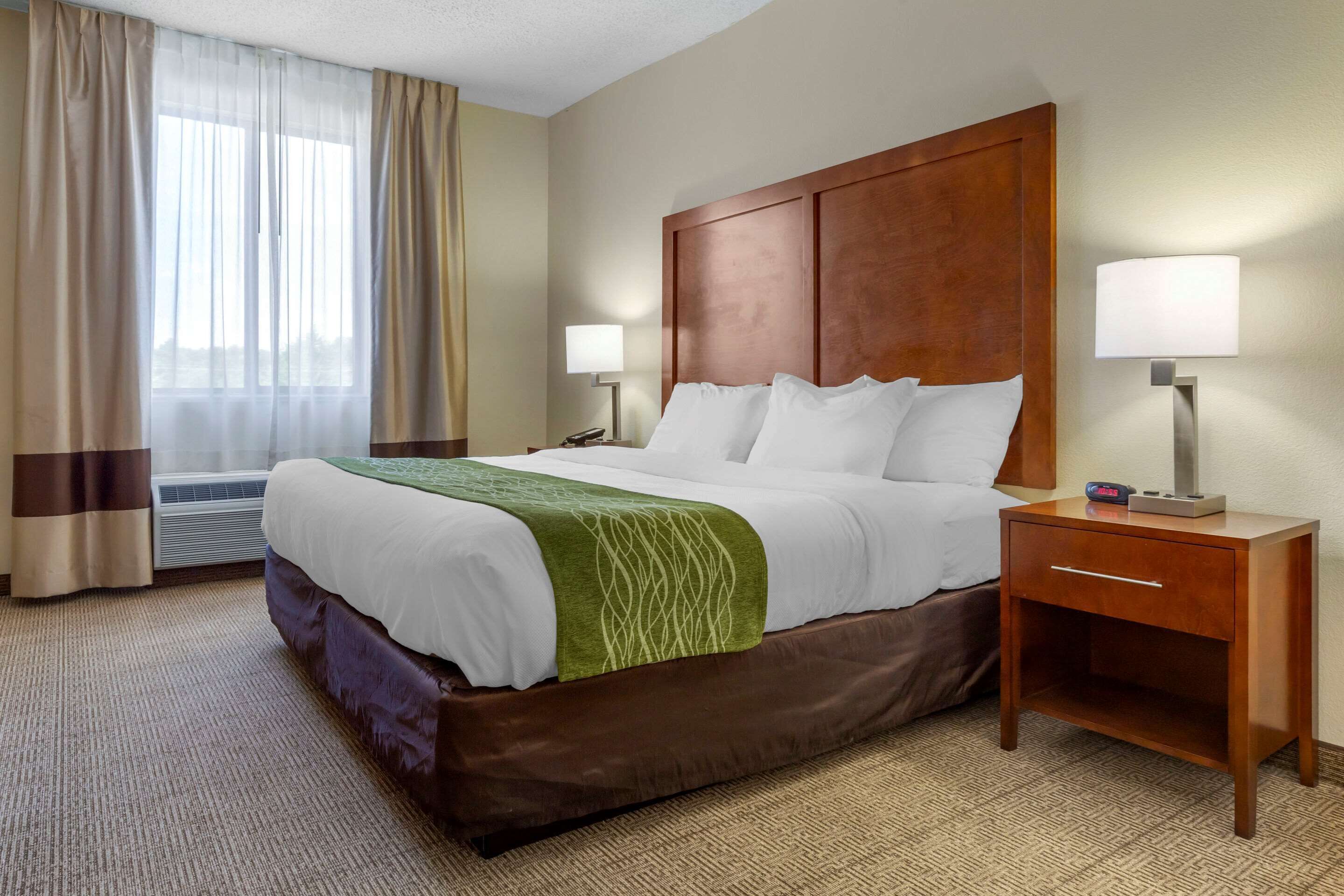 Una habitación de hotel con una cama grande, dos mesitas de noche, una lámpara y un ventanal con cortinas.