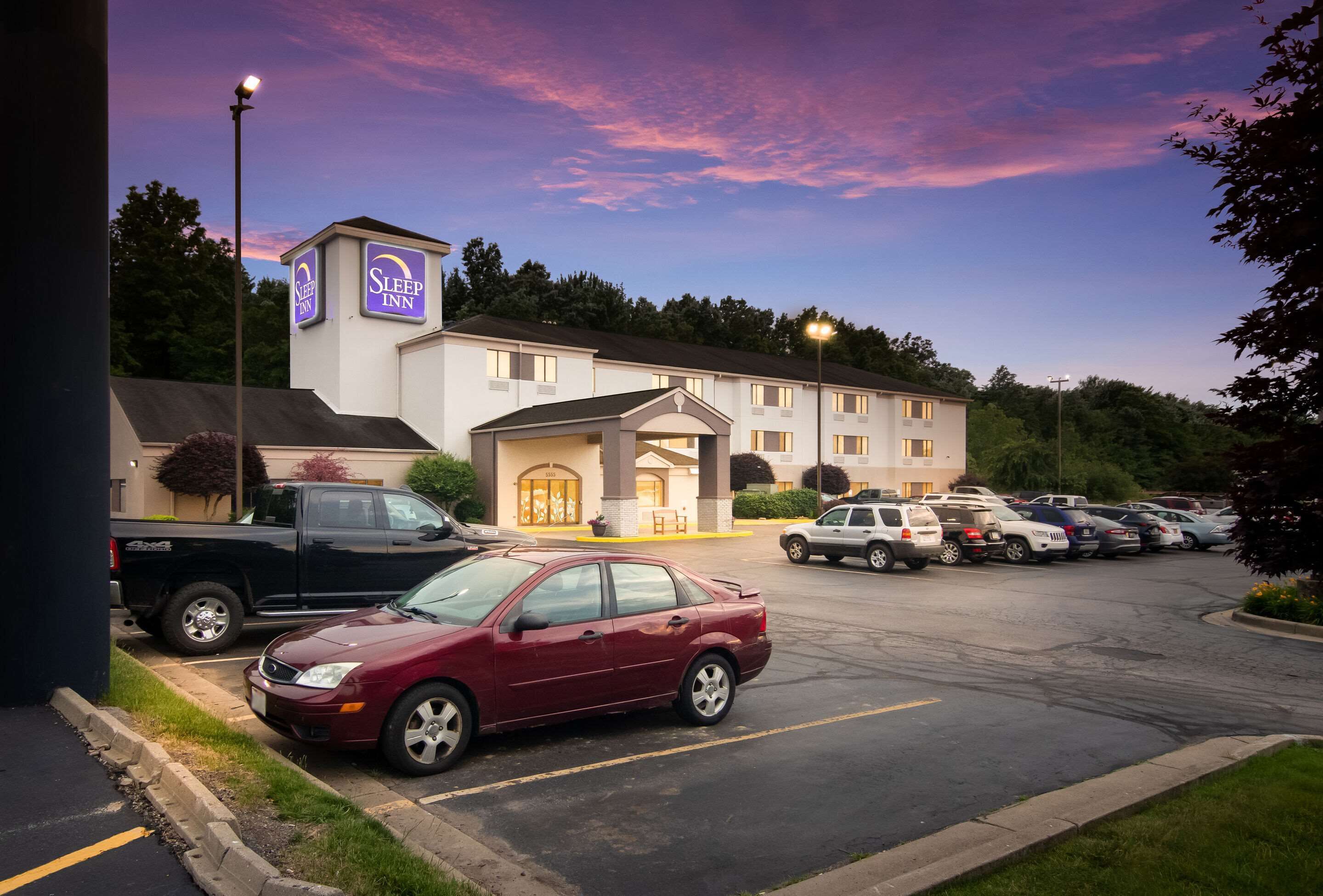 Un hotel Sleep Inn con un estacionamiento, un coche rojo estacionado al frente y un cielo despejado.