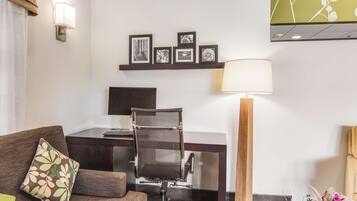 A hotel room with a desk, a chair, a lamp, a sofa with a floral pillow, and a wall with framed pictures.