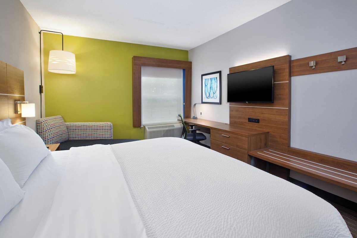 A hotel room with a large bed, a desk with a chair, a flat-screen TV mounted on the wall, and a window with blinds.