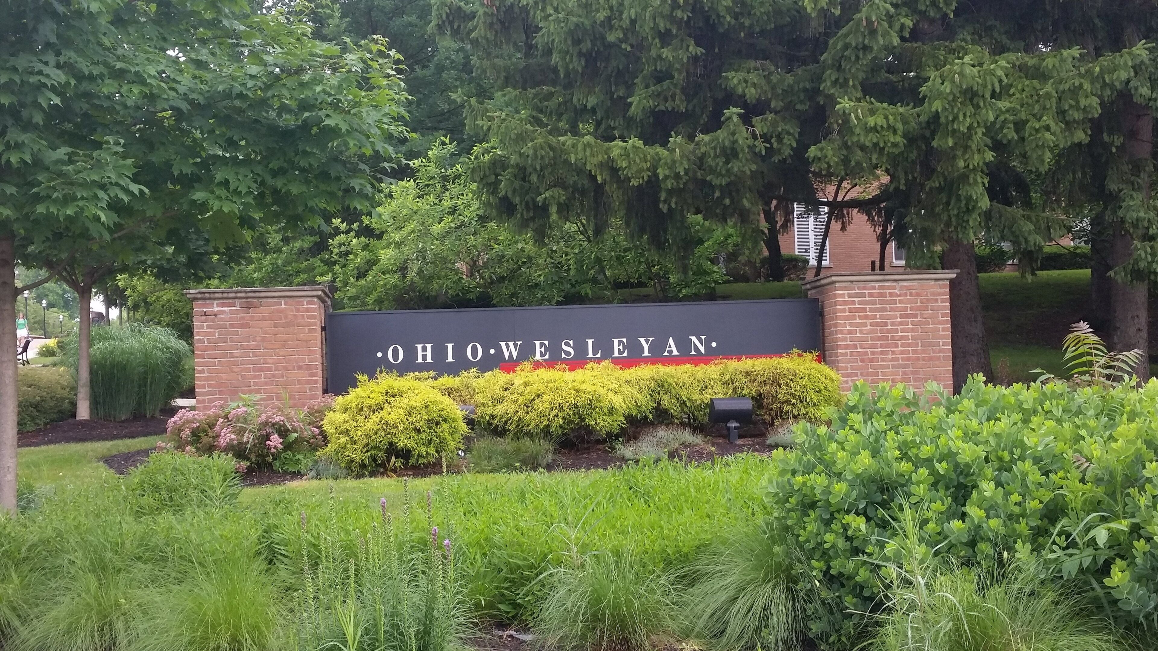 Ein Schild mit der Aufschrift 'OHIO WESLEYAN', umgeben von Grünpflanzen und Bäumen.