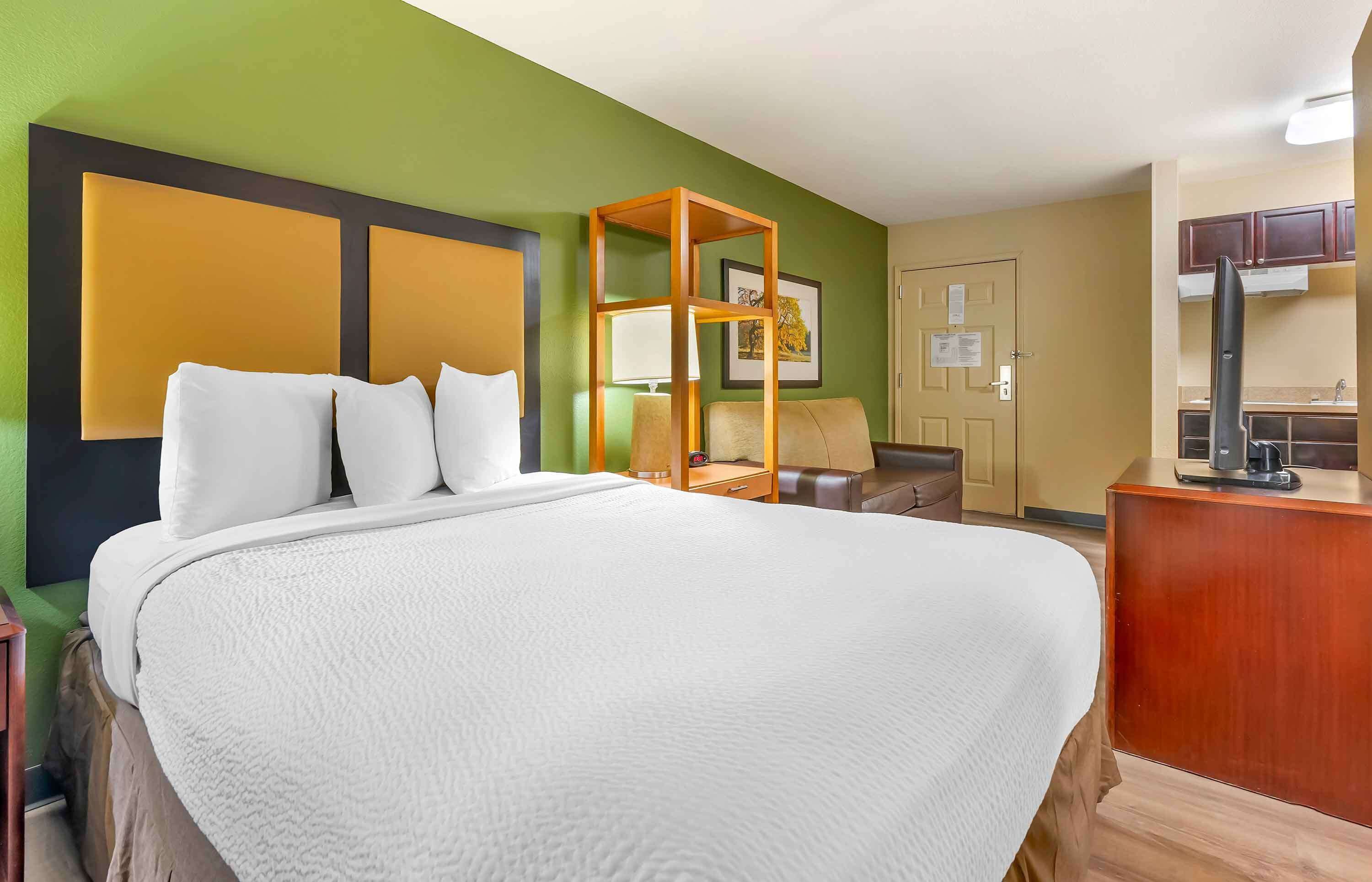 A hotel room with a large bed, a wooden headboard, a desk with a chair, a small table, and a kitchenette area.