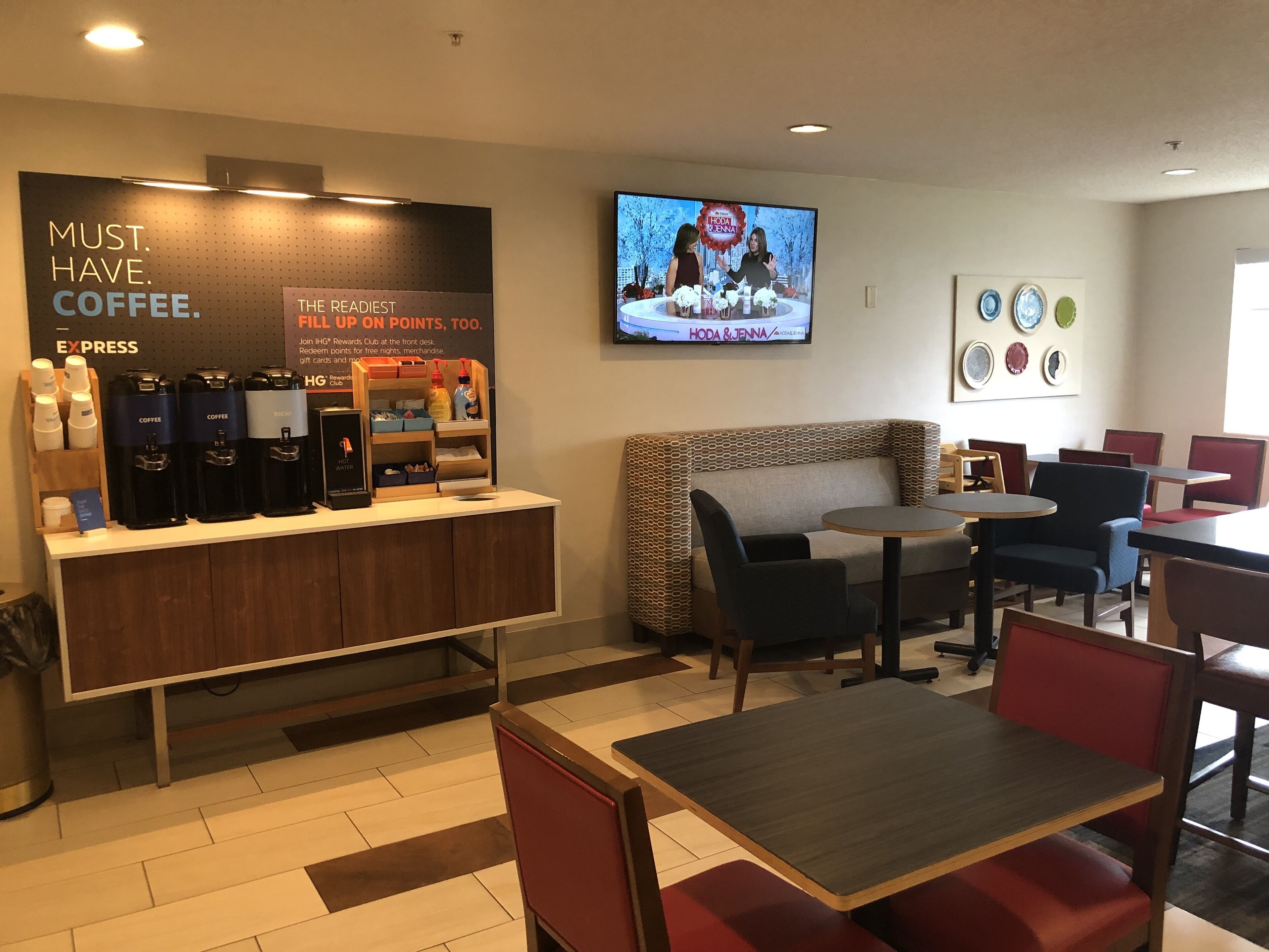 Un espace dédié au café, équipé de plusieurs distributeurs de café et d’une affiche faisant la promotion d’un programme de fidélité. On y trouve également un écran de télévision diffusant un programme, un coin salon avec des tables et des chaises, ainsi qu’un mur décoré d’assiettes.