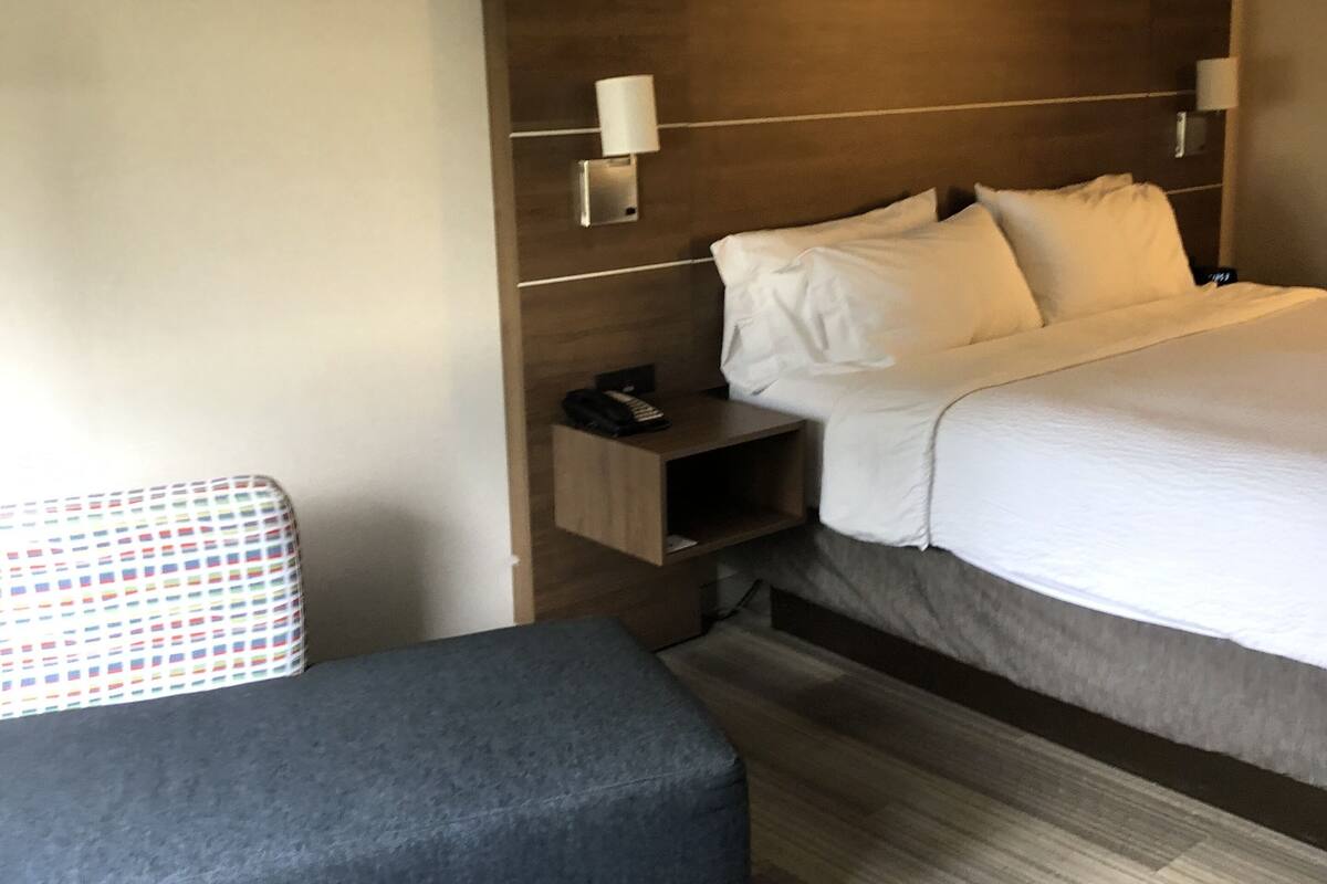 A hotel room with a large bed, a bedside table with a lamp, a small bedside table with a phone, and a grey bench.