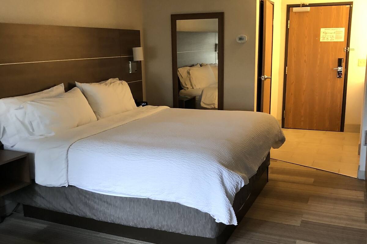 A hotel room with a large bed, a bedside table, a nightstand, a mirror, a door, and a light fixture on the ceiling.