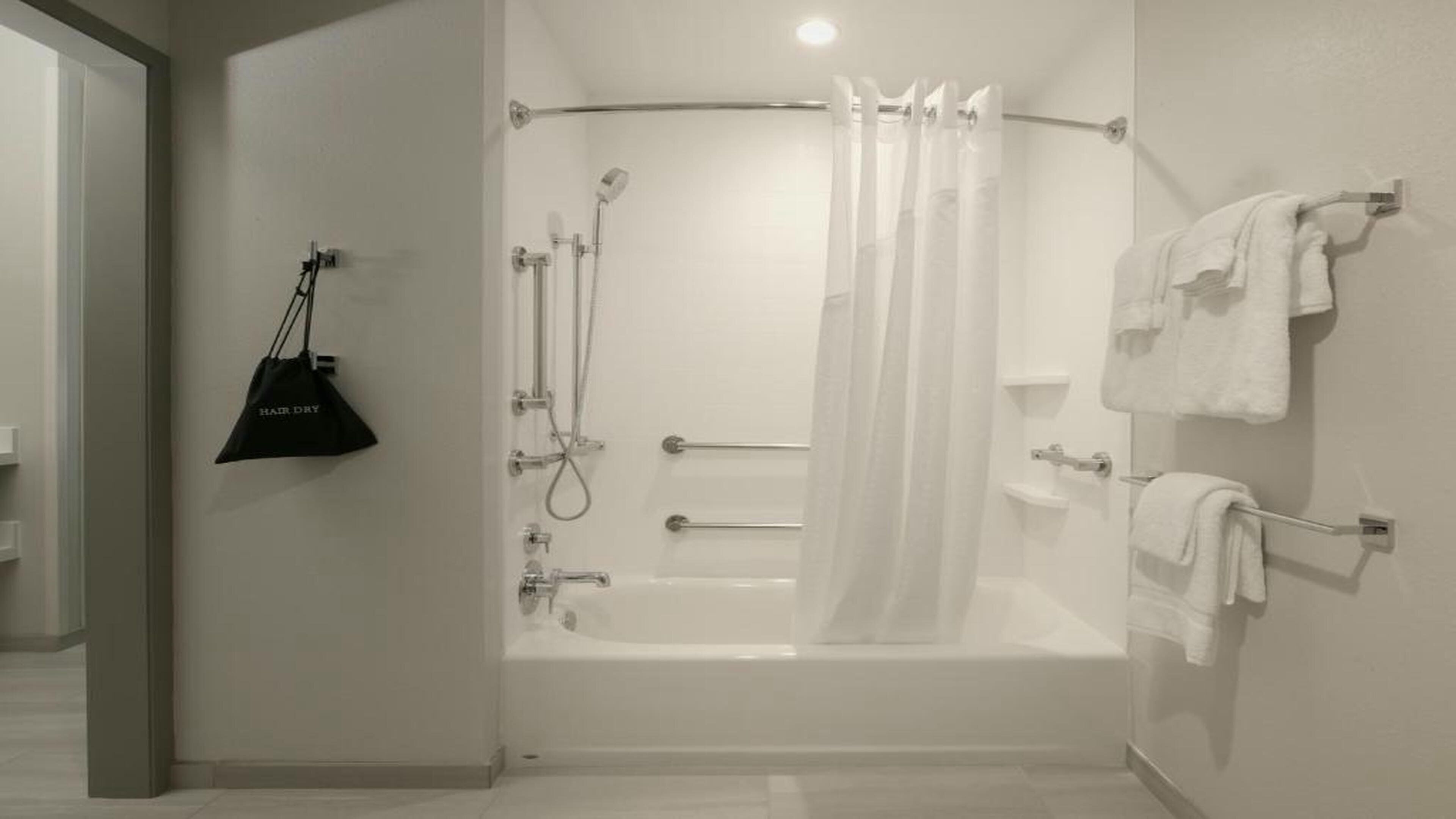 A modern bathroom with a bathtub, shower, and toilet. There is a towel rack with towels and a black bag hanging on the wall.