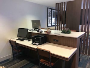 A hotel room with a desk, two chairs, a laptop, a desktop computer, a printer, and a decorative plant.