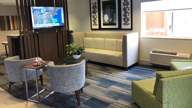A modern hotel lobby with a sofa, chairs, a small table with a plant, a TV displaying a weather forecast, and framed artwork on the wall.