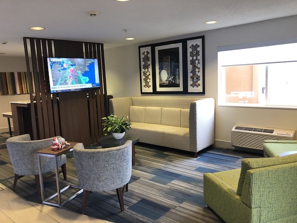 A modern hotel lobby with a sofa, chairs, a small table with a plant, a TV displaying a weather forecast, and framed artwork on the wall.