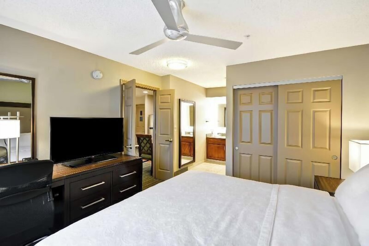 Une chambre d’hôtel avec un lit, un téléviseur posé sur une commode, un ventilateur de plafond et une porte donnant sur une salle de bain.